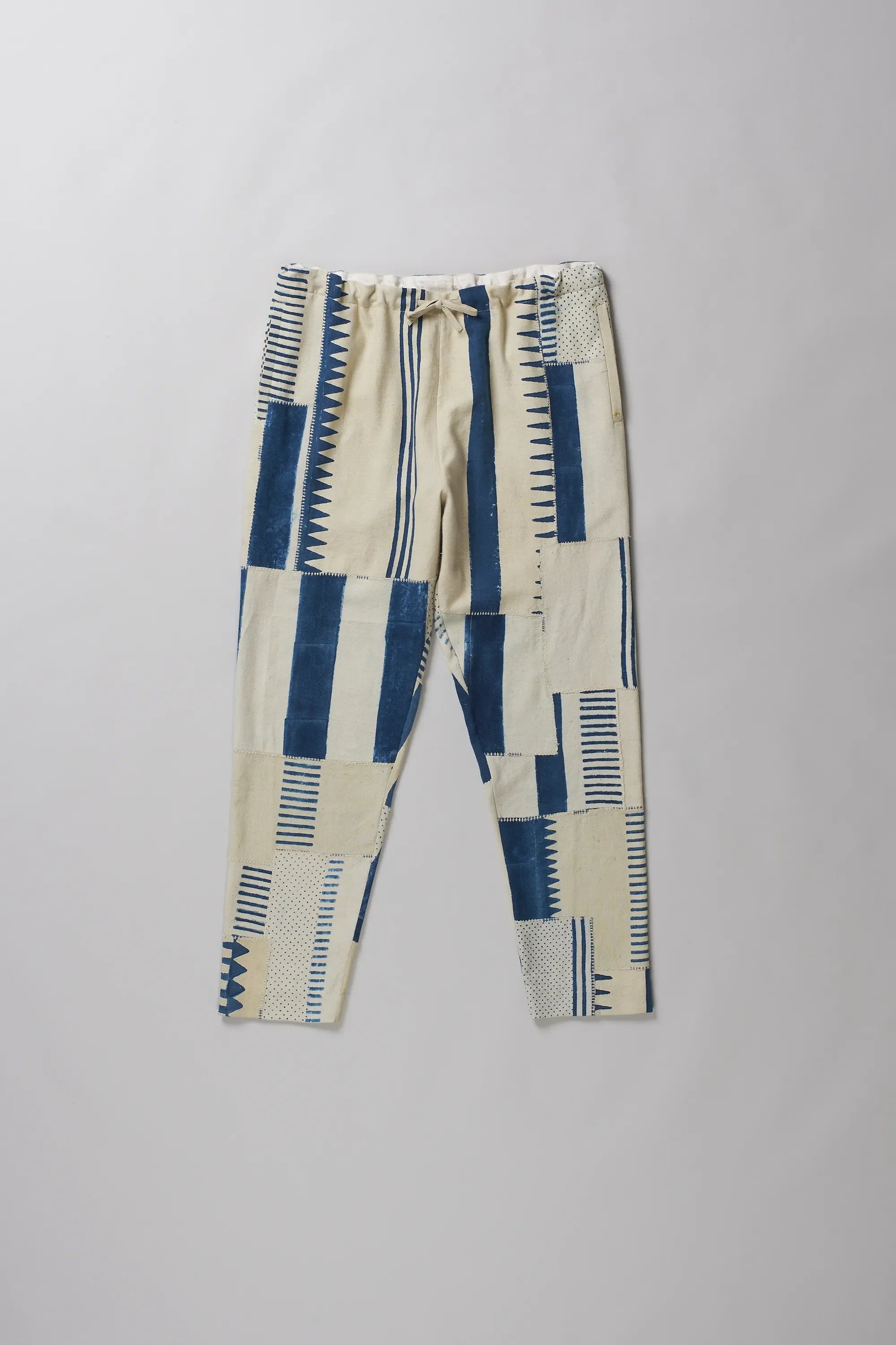 A pants with a geometric patchwork pattern in blue and cream, featuring a drawstring waist, laid flat on a light grey background.