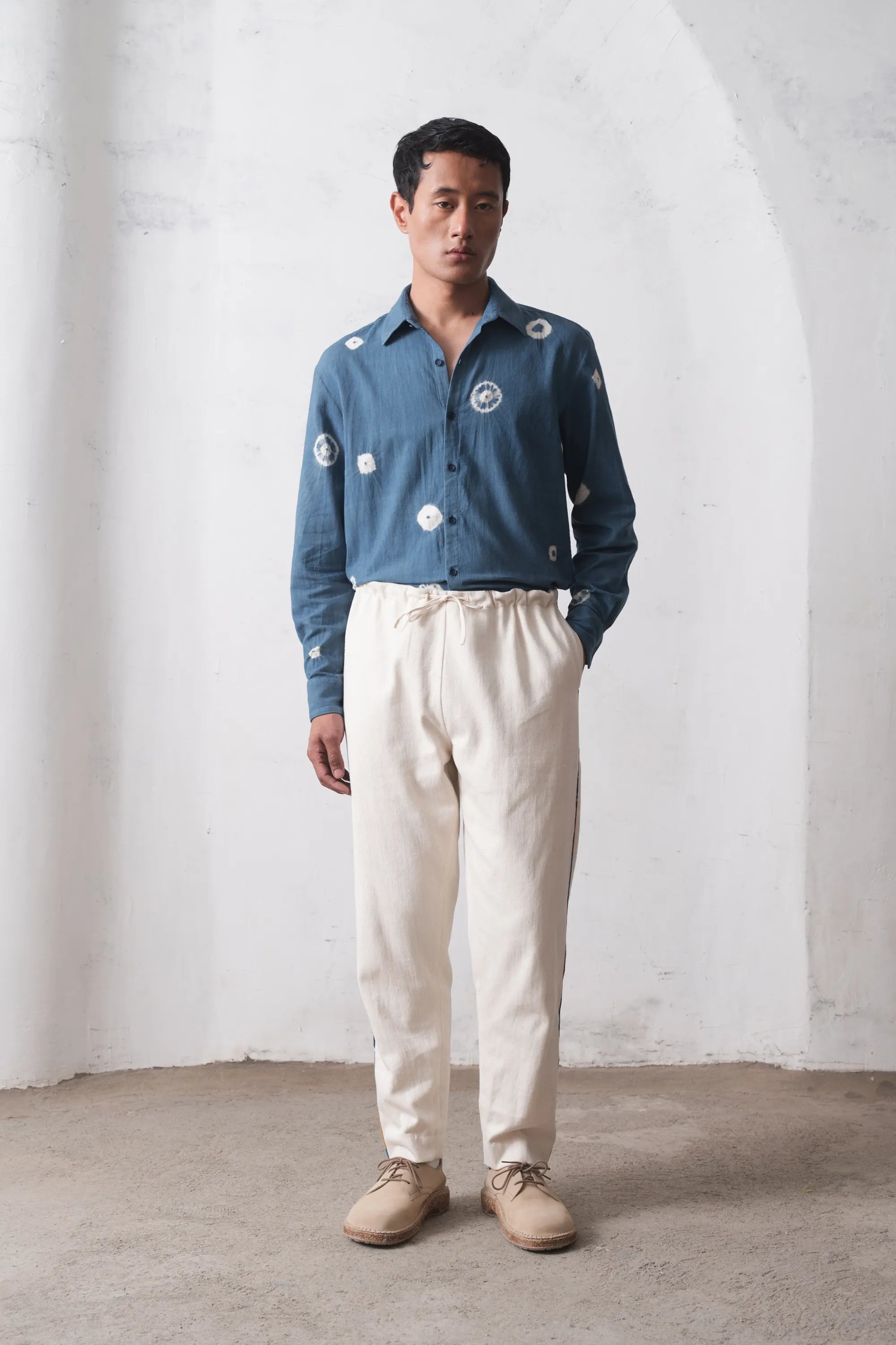 Man wearing a blue shibori shirt and white pants standing against a white wall