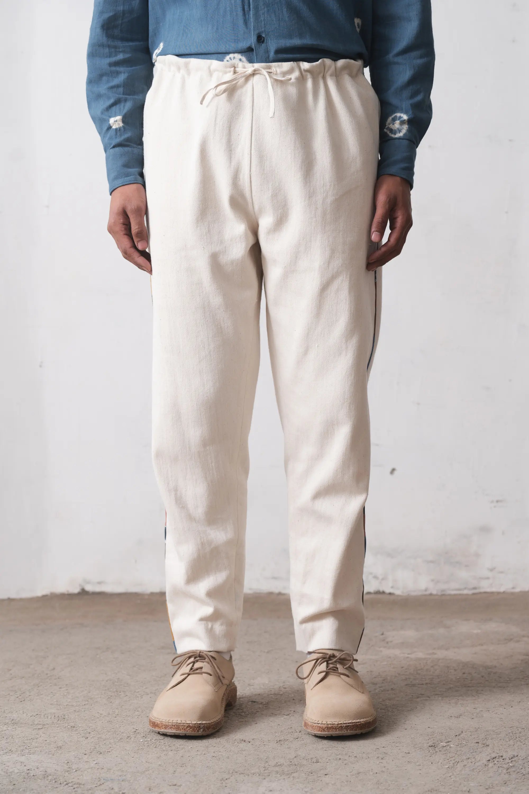 Person wearing ecru pants and a blue shirt against a white wall